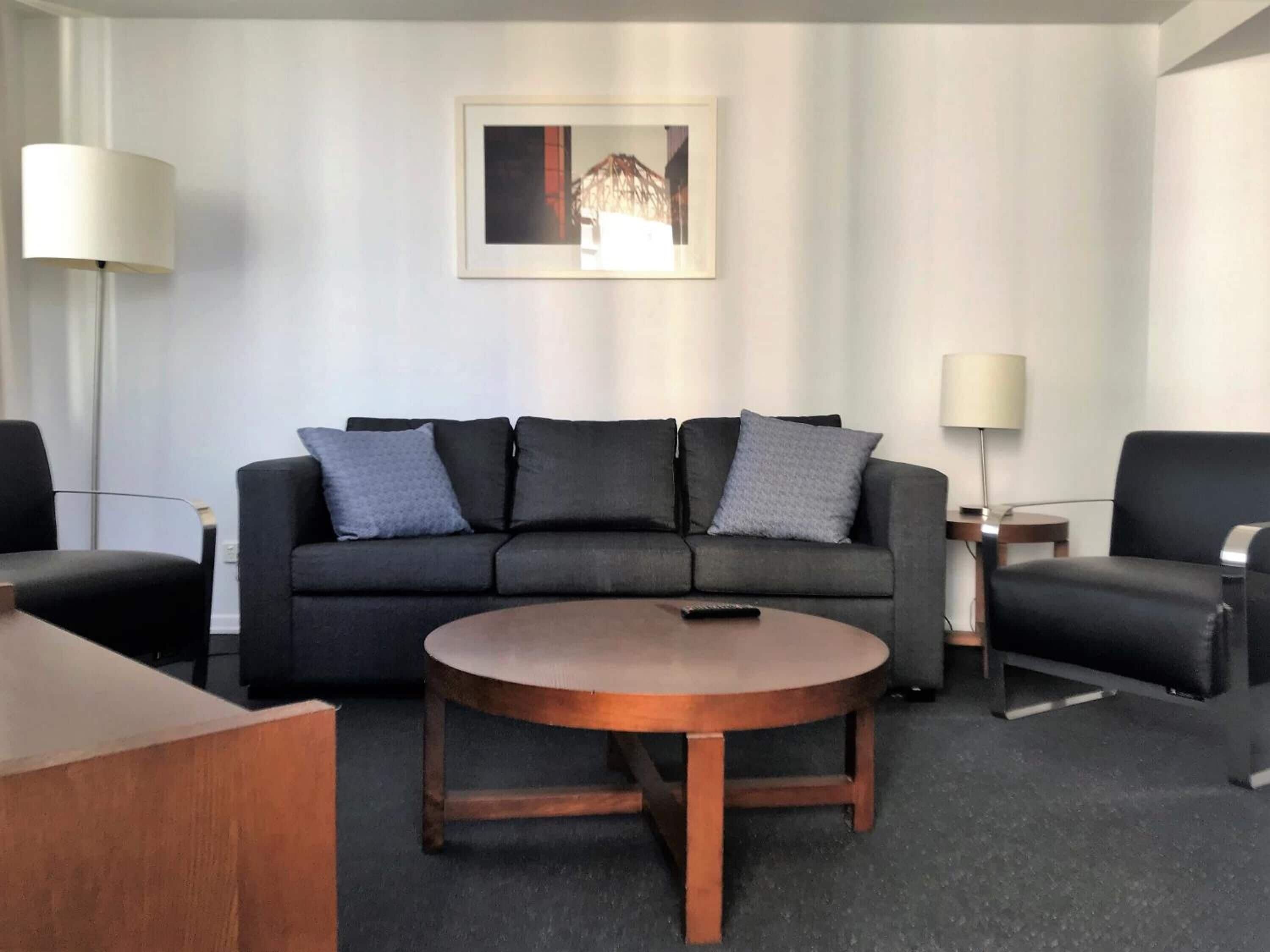 A modern hotel room with a black sofa, a round wooden coffee table, and two black armchairs.