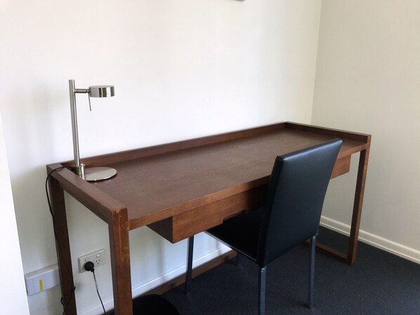 A wooden desk with a black chair, a lamp, and a power outlet on the wall.