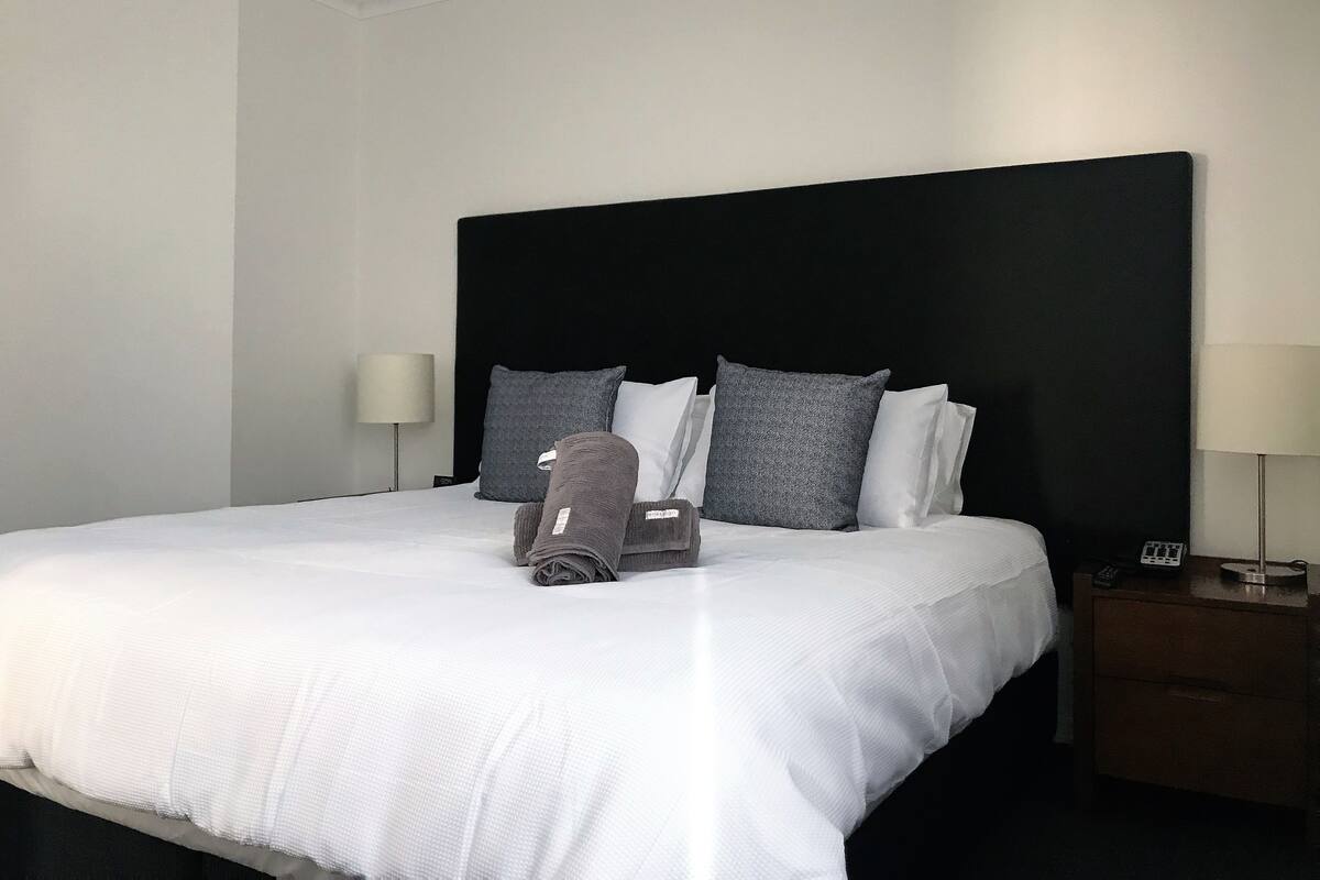 Un lit bien fait, avec une literie blanche, une tête de lit noire et plusieurs oreillers dans différentes nuances de gris et de blanc. Une serviette pliée est posée sur le lit, et une table de chevet est équipée d’une lampe et d’une télécommande.