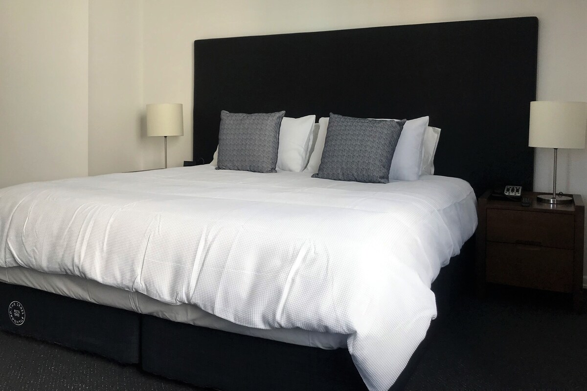 Uma cama bem arrumada com cabeceira preta, roupa de cama branca e duas mesas de cabeceira com abajures.