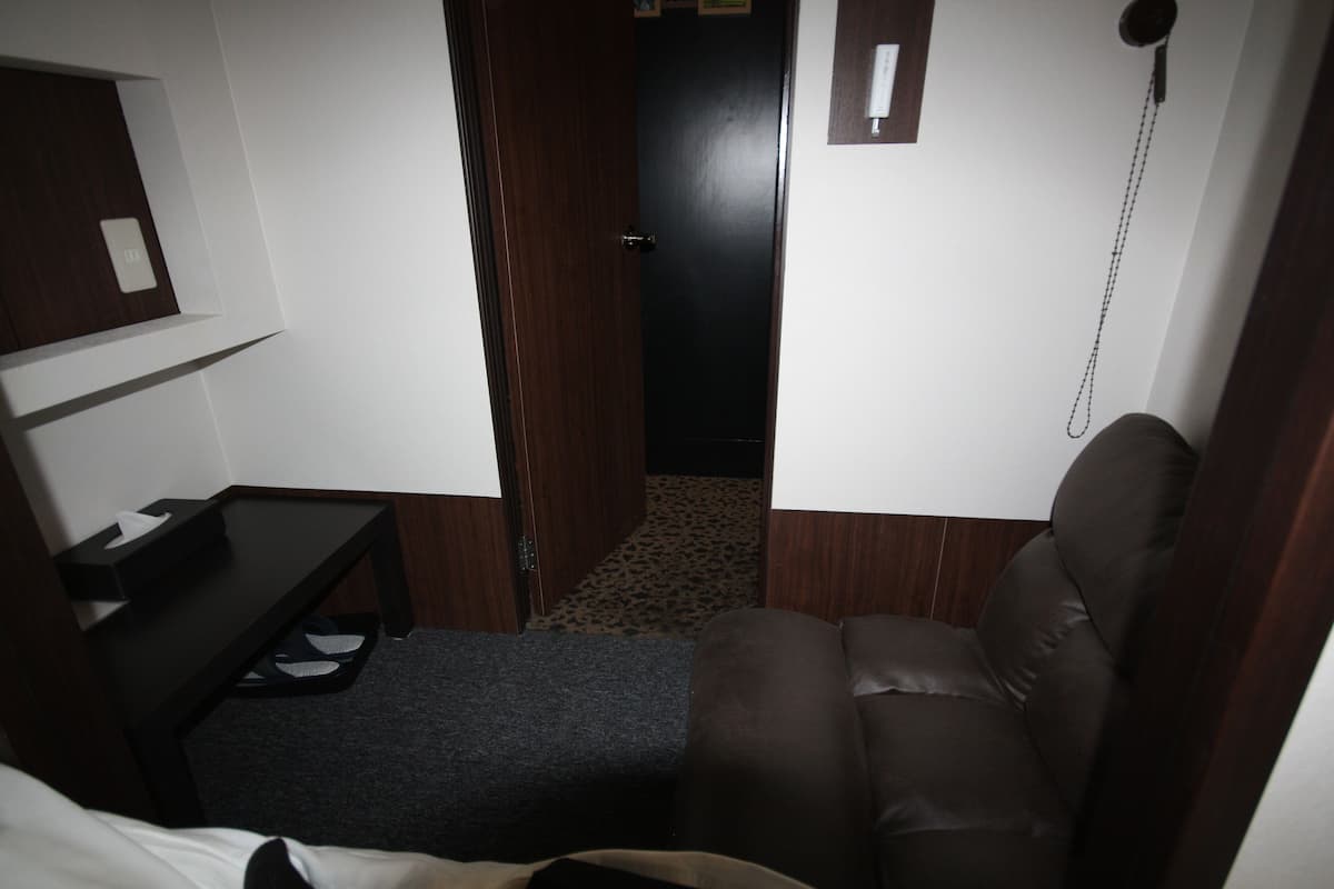 A small, dimly lit room with a single bed, a small table with a tissue box, a brown leather chair, and a door leading to another room.