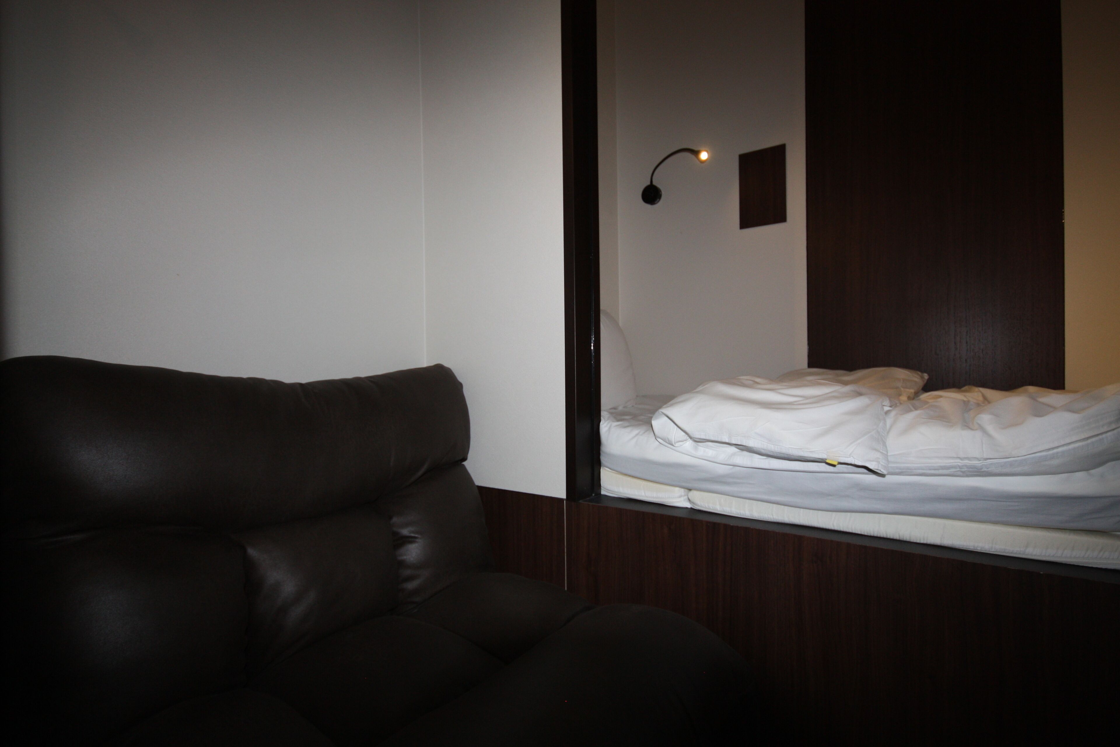 A room with a dark brown leather sofa, a white bed with a white mattress, and a wall-mounted light fixture.