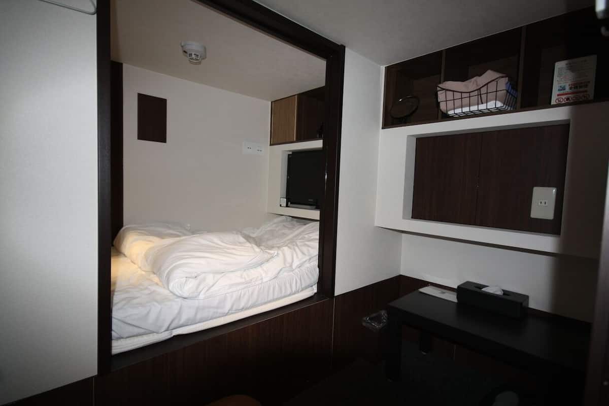 A compact hotel room with a bed, a small desk, a shelf with a basket, and a wall-mounted TV.
