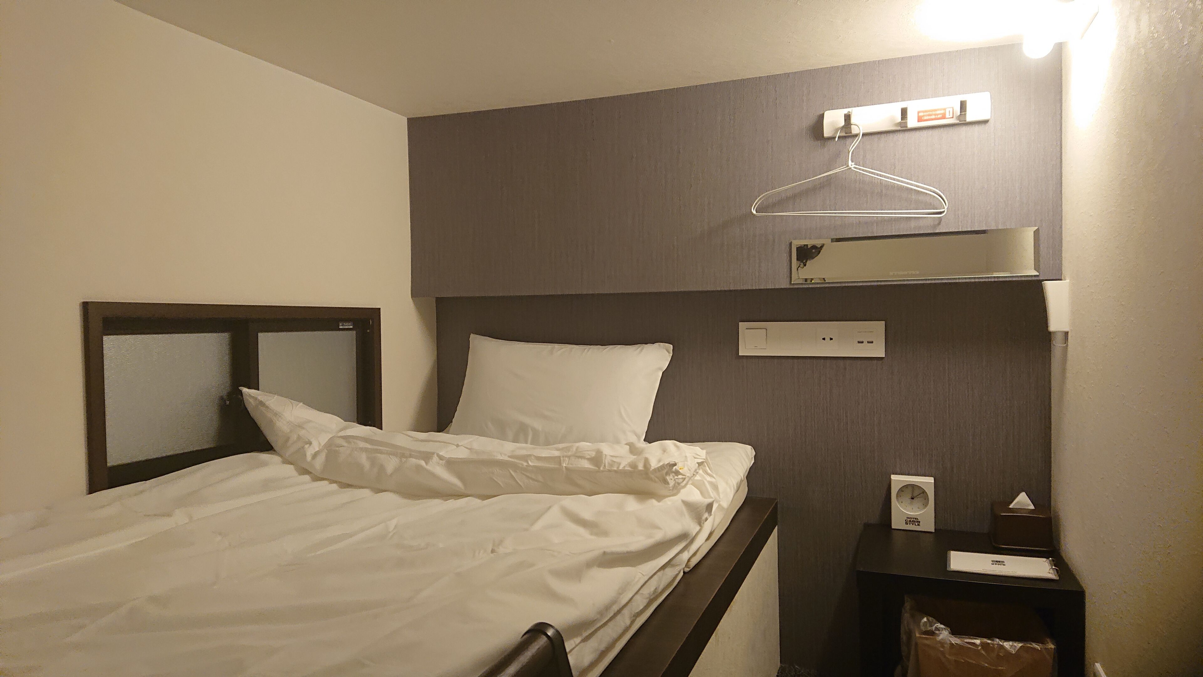 Une chambre d’hôtel avec un lit, une table de chevet avec une horloge et une boîte de mouchoirs, un porte-manteau mural et un lampadaire fixé au mur.