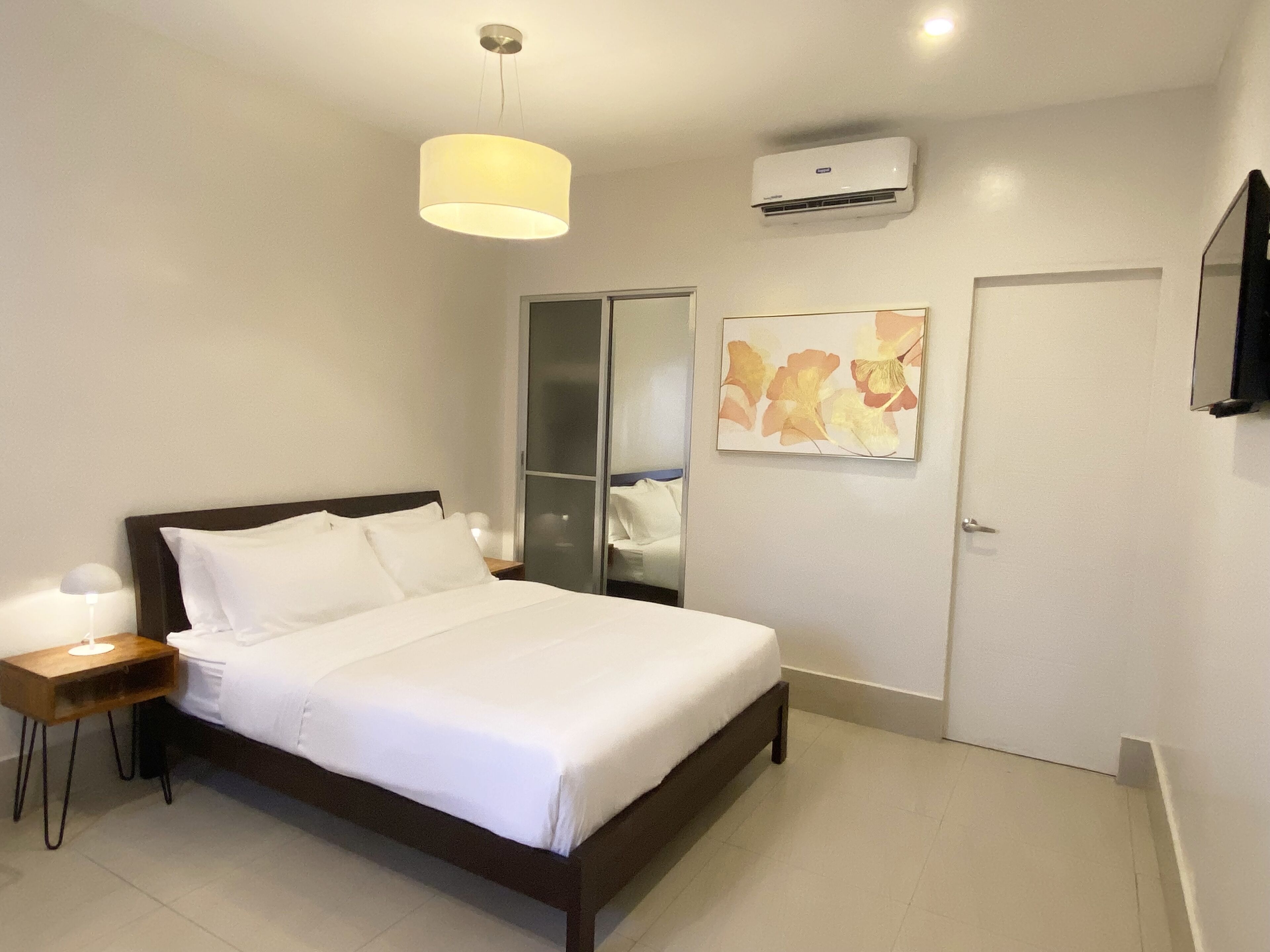 Un lit bien fait dans une chambre d’hôtel, avec une table de chevet, un climatiseur fixé au mur et une porte dotée d’un miroir.