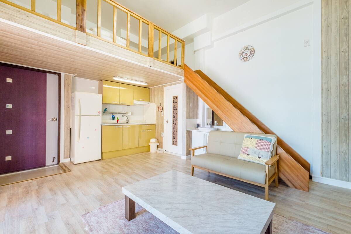 Amplia sala de estar con techo alto, escalera de madera, cocina y un sofá con un cojín decorativo.