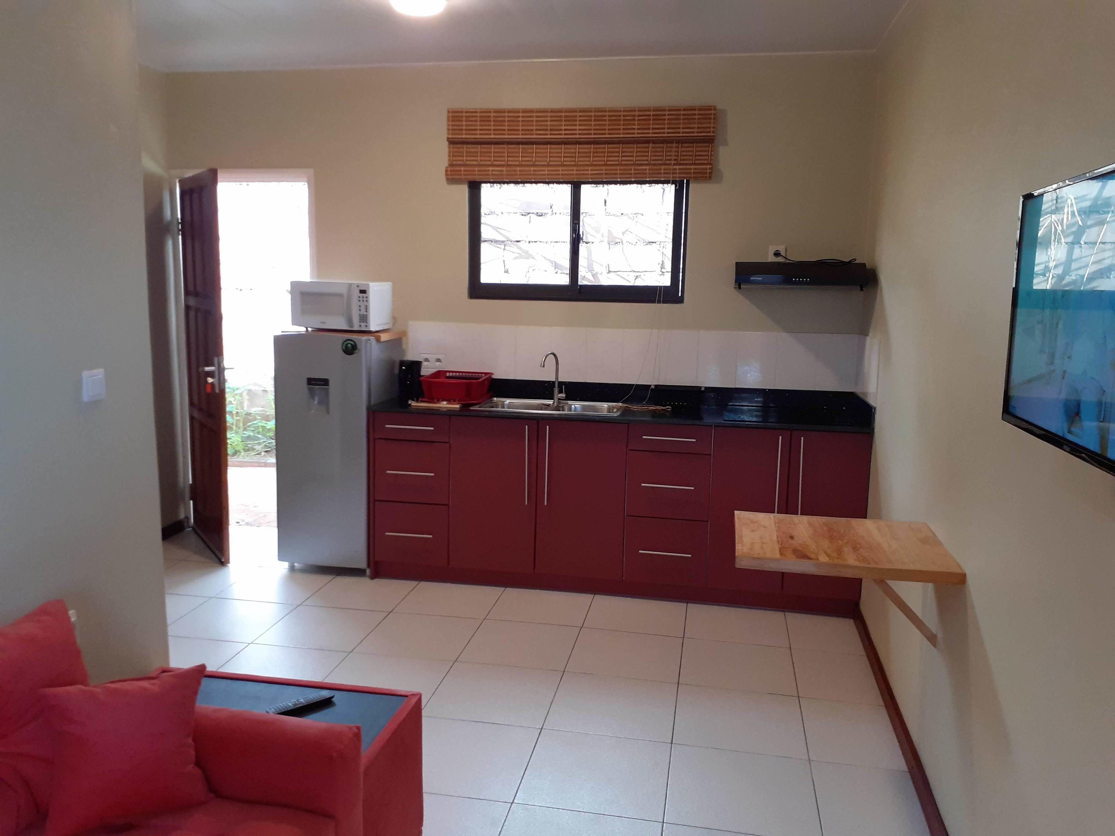 A compact kitchen with a refrigerator, microwave, and a window with blinds. There is a red sofa and a small table with a remote control.