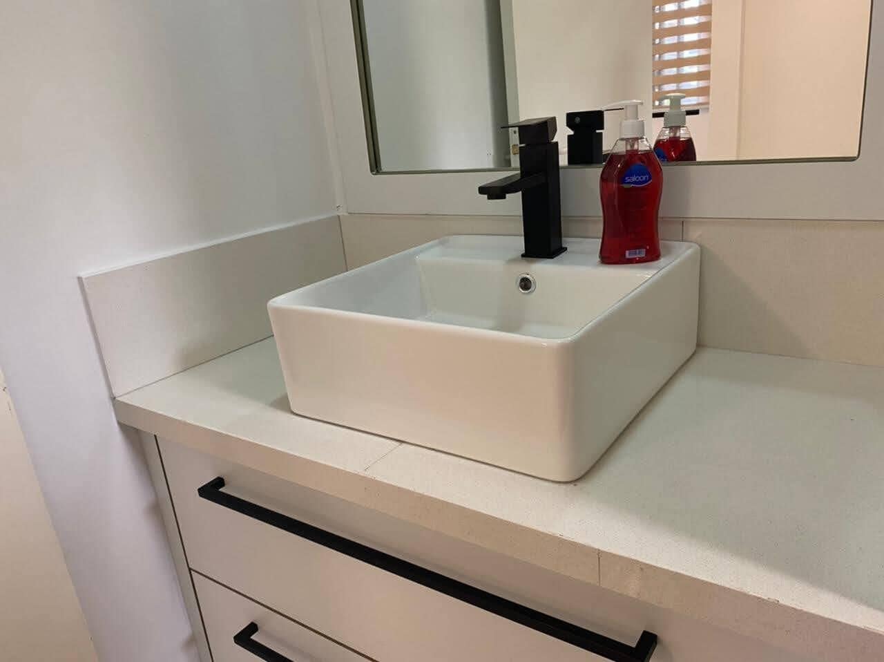 Une salle de bain blanche avec un lavabo rectangulaire, un robinet noir et deux bouteilles de savon rouge posées sur le comptoir.