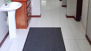 A hallway with a white tiled floor, a black mat at the entrance, a wooden cabinet with a sink, and a door leading to another room.