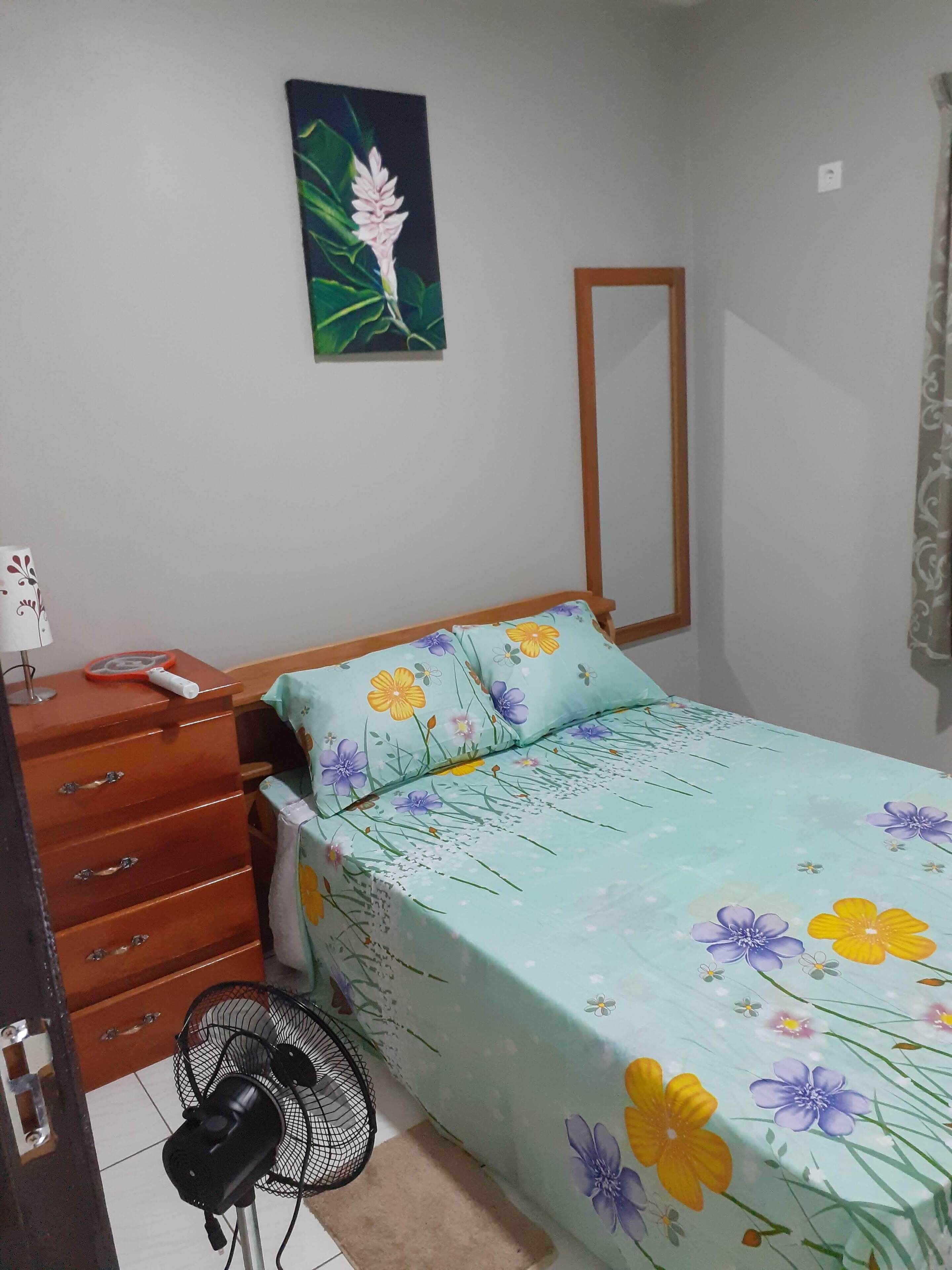 A bedroom with a bed, a dresser, a mirror, a fan, and a wall-mounted painting.