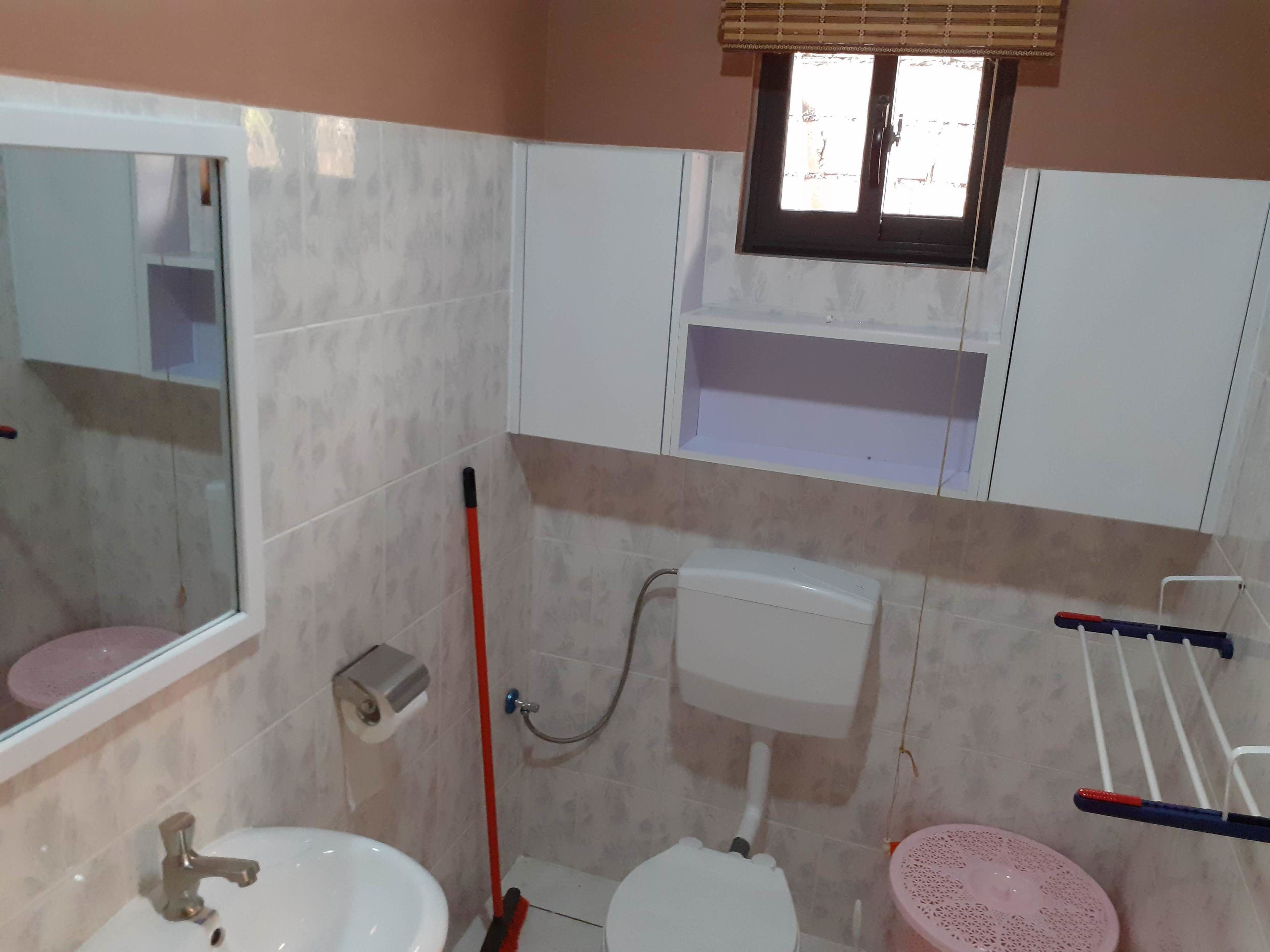 A bathroom with a toilet, sink, and shower. There is a window above the toilet and a mirror on the wall.