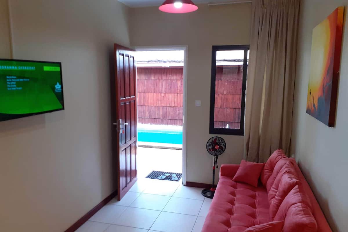 A living room with a red sofa, a television on the wall, a fan, and a view of a wooden fence outside.