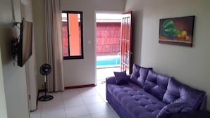 A living room with a purple sofa, a standing fan, a painting of fruits, and a view of a wooden deck and pool.