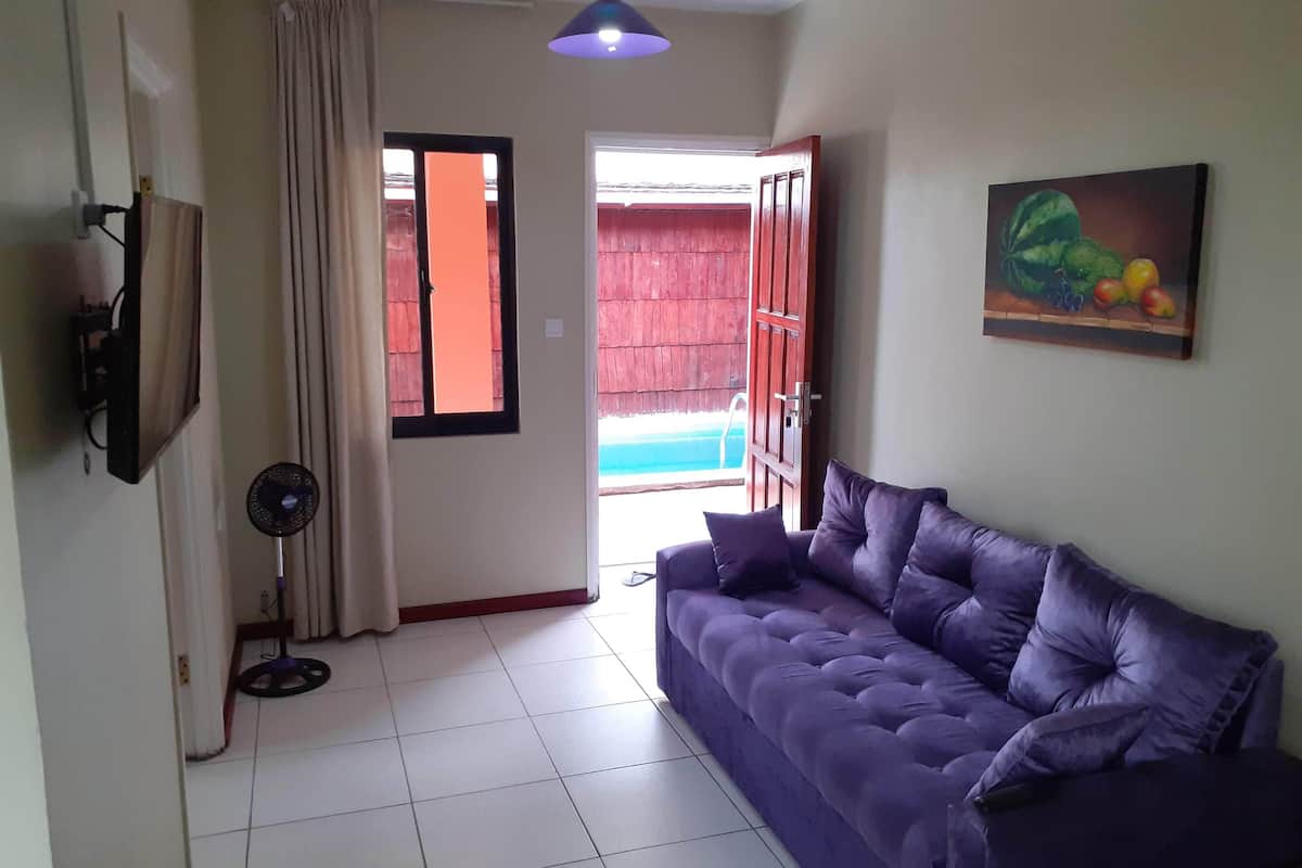 A living room with a purple sofa, a standing fan, a painting of fruits, and a view of a wooden deck and pool.