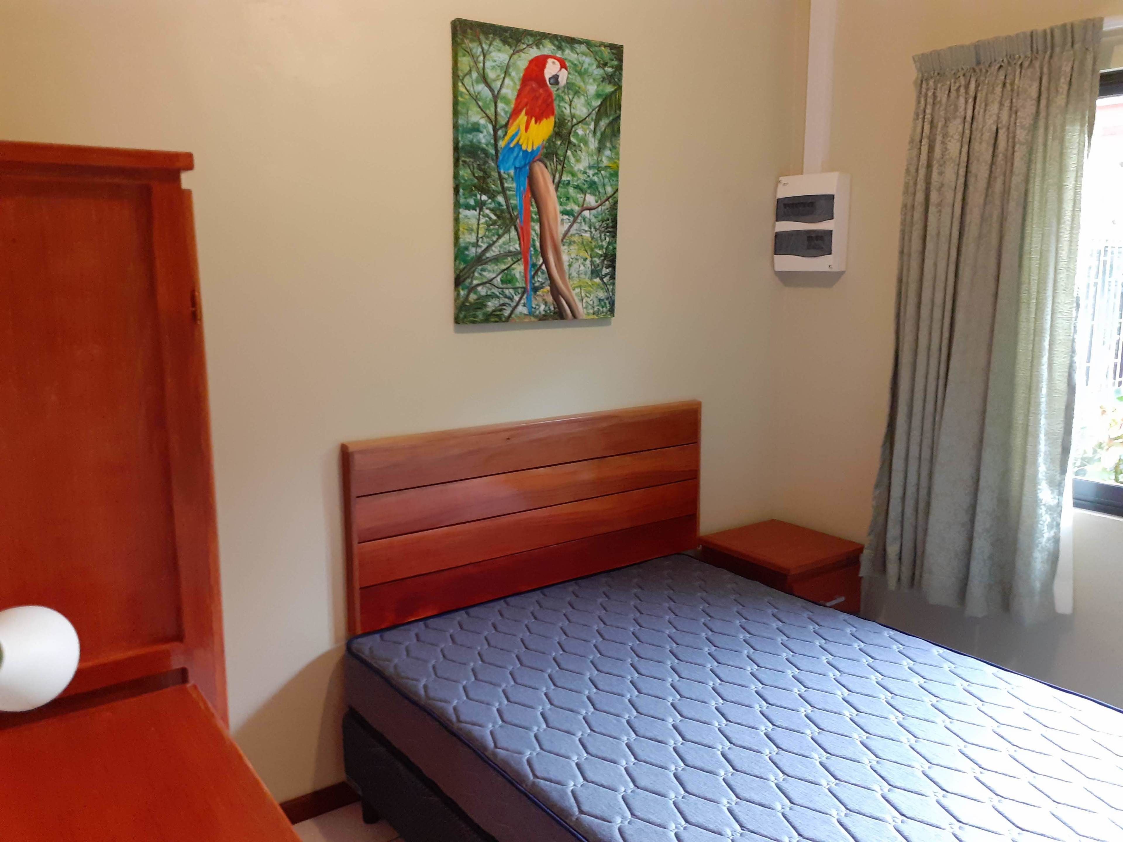 Un dormitorio con cabecera de madera, colchón azul, mesita de noche pequeña, aire acondicionado de pared y una ventana con cortinas.