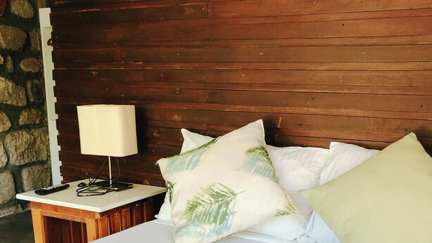 A neatly made bed with white linens, a basket of folded towels, and a nightstand with a lamp and remote control. - Ted's Bed and Breakfast (Santa Cruz)