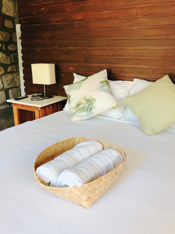 A neatly made bed with white linens, a basket of folded towels, and a nightstand with a lamp and remote control. - Ted's Bed and Breakfast (Santa Cruz)
