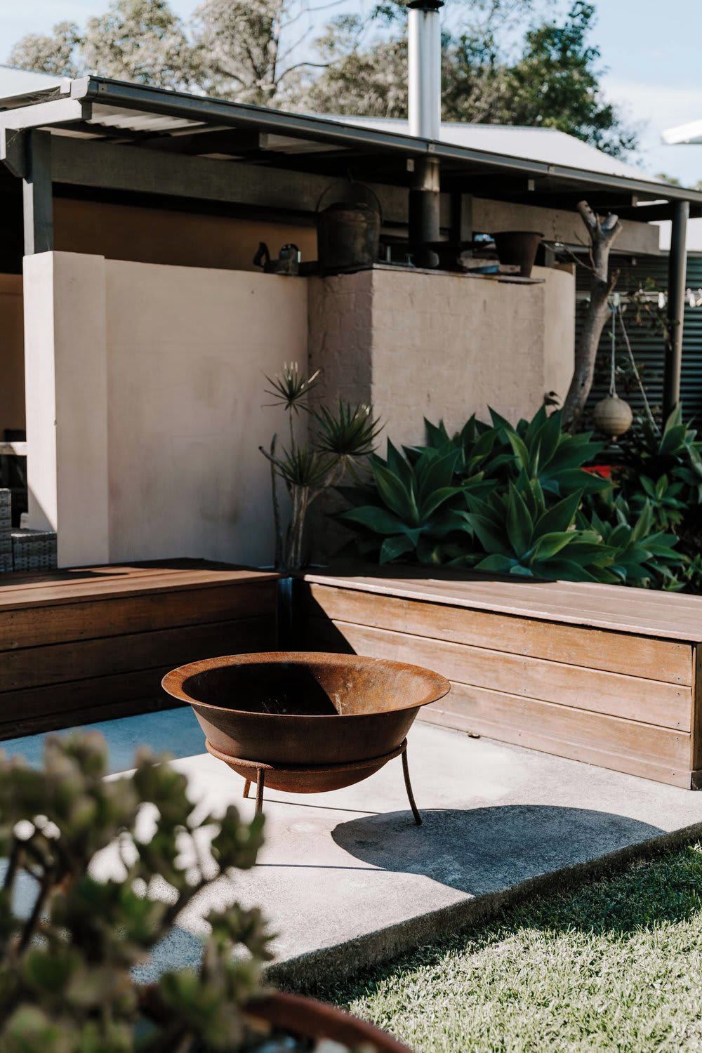 A modern outdoor space with a concrete structure, a metal fire pit, and various plants.