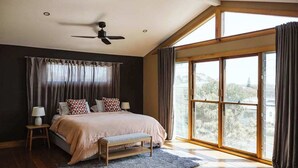 Une chambre avec un grand lit, un ventilateur de plafond, une table de chevet, une lampe, un banc et une vue dégagée grâce à de grandes fenêtres.