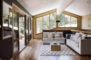 A spacious living room with a large sofa, a coffee table, and a view of the outdoors through large windows. - The Agave (Currarong)