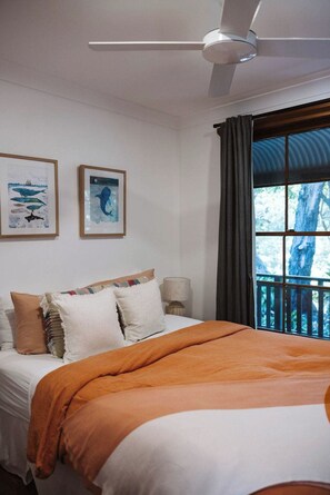 A bedroom with a bed, a ceiling fan, two framed pictures, a window with curtains, and a view of trees.