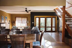 A spacious living area with a dining table, chairs, and a sofa. There is a staircase on the right and a large sliding glass door leading outside. - The Agave (Currarong)