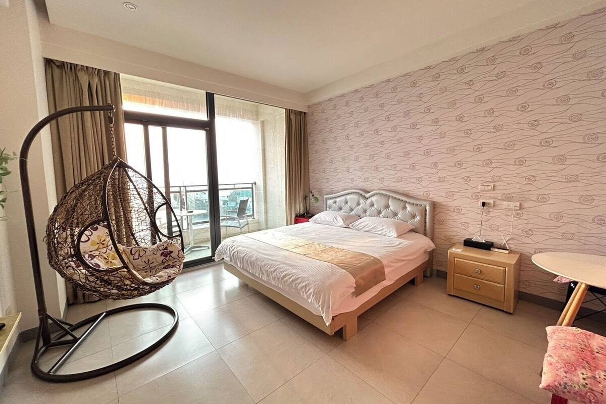 A modern hotel room with a large bed, a hanging chair, a small table, and a view of the city through the window.
