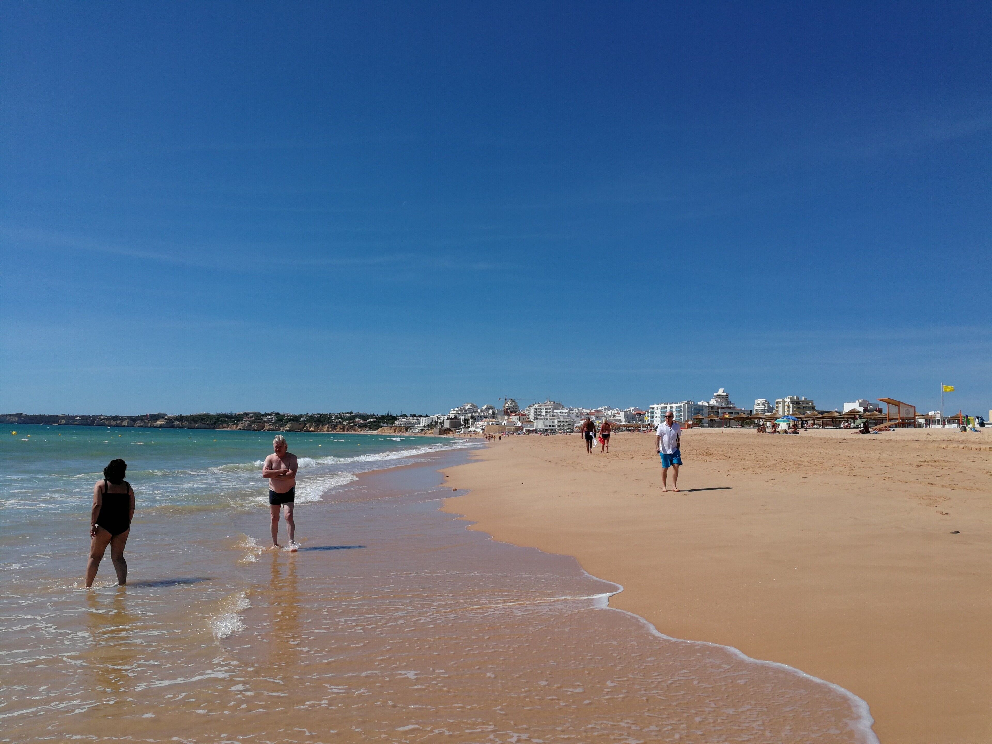 Een strand met mensen die langs de kust wandelen, een heldere lucht en gebouwen op de achtergrond.