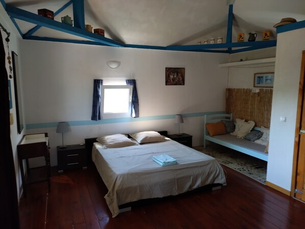 A bedroom with a large bed, a smaller bed, a desk, and a chair. There is a window with curtains, a wall-mounted shelf with decorative items, and a framed picture on the wall.