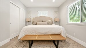 A bedroom with a bed, bedside lamps, a bench, a window with blinds, and a patterned rug. - Bright 3BR · 5 Mi to Disneyland · Sleeps 8, 2 BA (Fullerton)