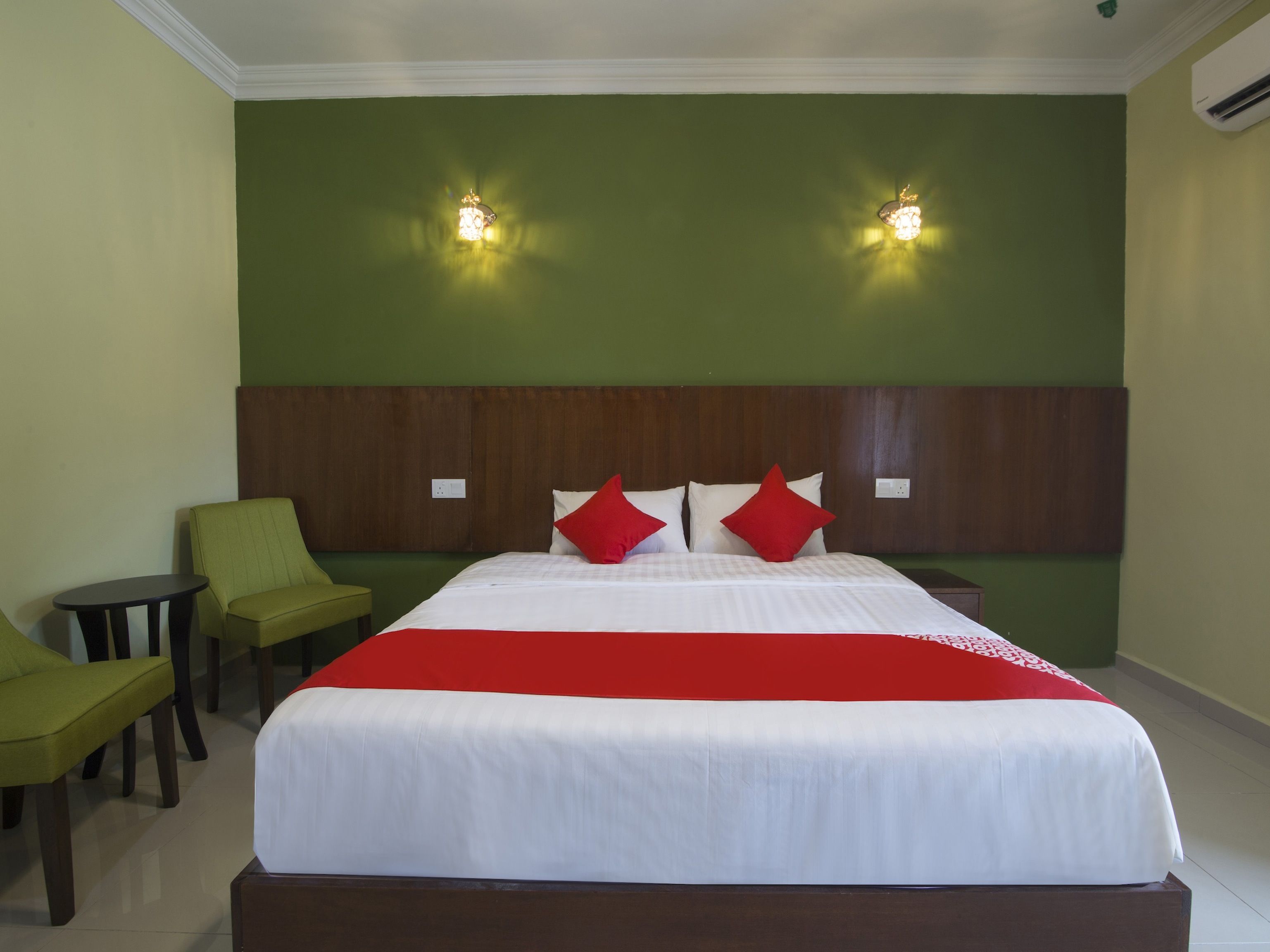 A hotel room with a large bed, green walls, two wall-mounted lights, a wooden headboard, and a green armchair.