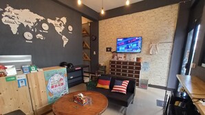 Un área de descanso moderna con una mesa redonda de madera, un sofá negro con un cojín con la bandera estadounidense, un mapa del mundo en la pared y un televisor mostrando información del mercado de valores.