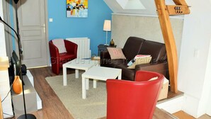 Un salon chaleureux avec un canapé rouge, une table basse blanche et un fauteuil rouge. Un tableau orne le mur et une lucarne se trouve au-dessus.