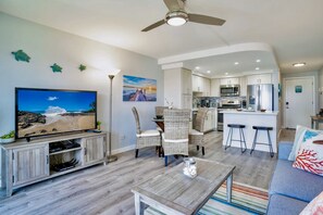 A modern living room with a flat-screen TV, wicker furniture, a ceiling fan, and a kitchen in the background. - NEW REMODELED, OCEAN VIEW, A/C IN ALL ROOMS, CAL KING BED, 4K, SMART TV, KEURIG (Kihei)