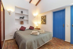 A bedroom with a bed, bedside lamps, a shelf with decorative items, a blue door, and a wall-mounted artwork.