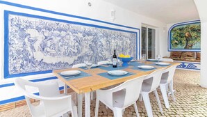 Een eetgedeelte met een grote blauw-witte betegelde muur, een houten tafel, witte plastic stoelen en een blauwe kom.