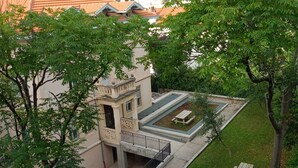 Un patio con un pequeño edificio, un área pavimentada con bancos y un camino bordeado de árboles.