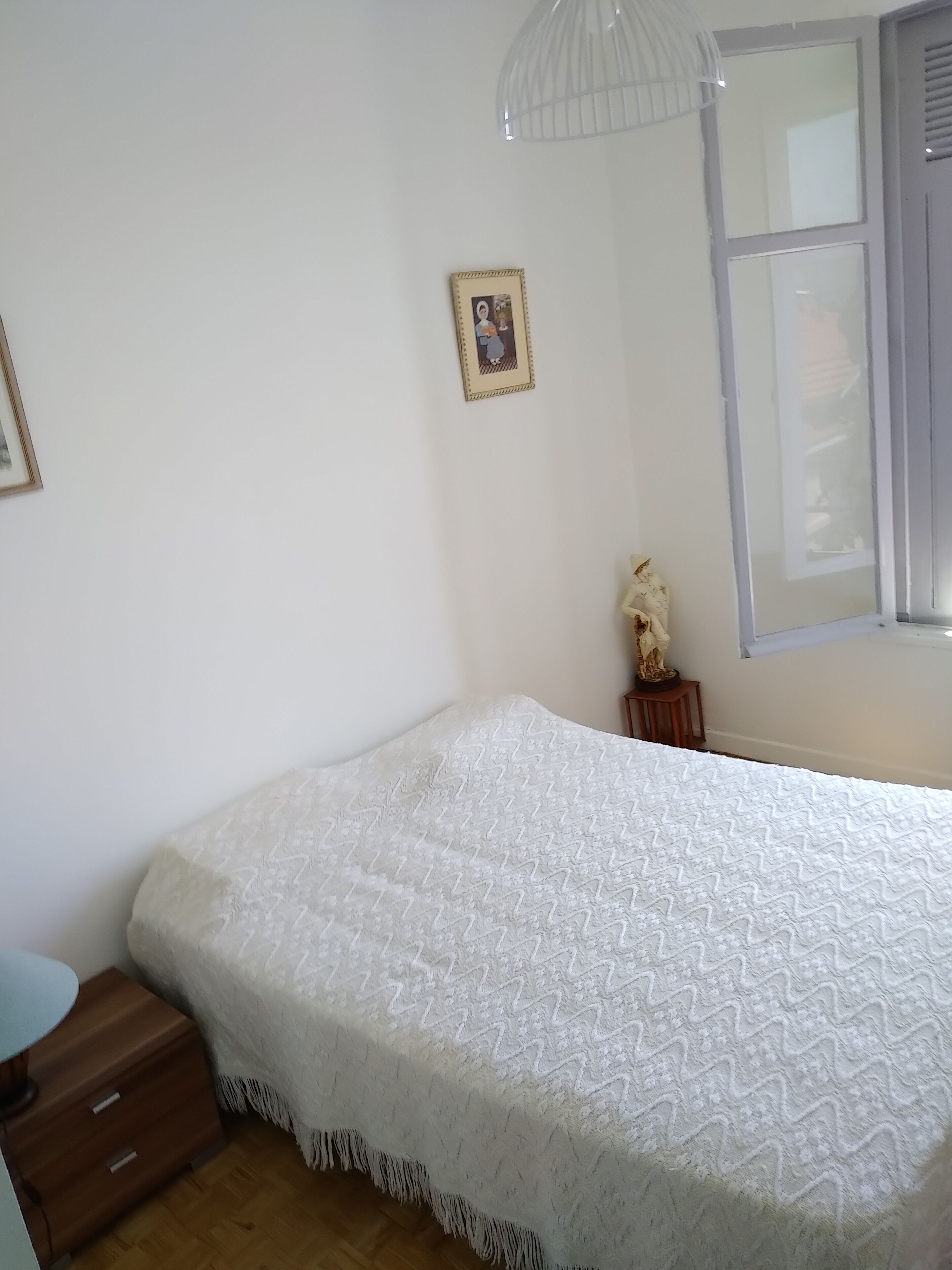A bedroom with a bed, a nightstand, a small statue, a framed picture, and a window with blinds.