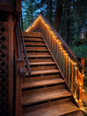A wooden staircase with railings and string lights, surrounded by trees.