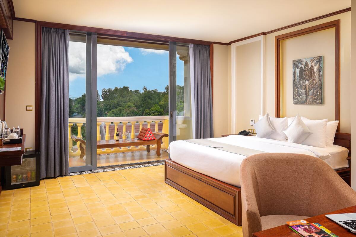 Habitación de hotel con una cama, un escritorio con una computadora portátil, una silla, un balcón con vistas a árboles y un cuadro en la pared.