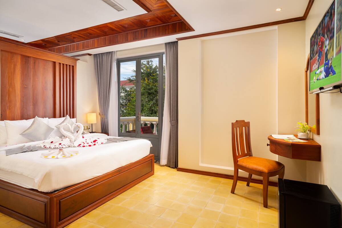 Habitación de hotel con una cama de madera, un escritorio con una silla, televisión y balcón con cortinas.