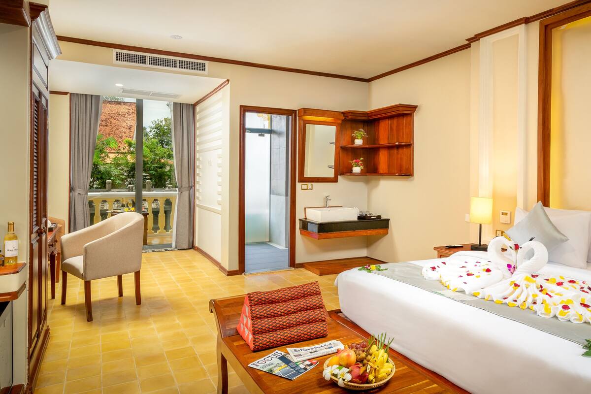 Habitación de hotel con cama, escritorio, silla, una pequeña mesa con frutero, revista, baño con lavamanos y espejo, y balcón con vistas a zonas verdes.
