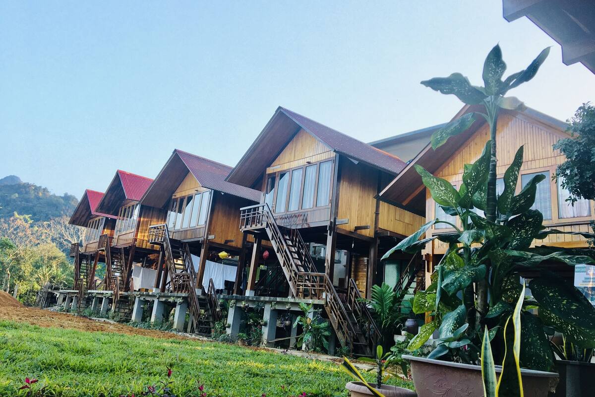A row of wooden cabins with balconies and stairs leading to the upper floors, surrounded by lush greenery and a clear sky.