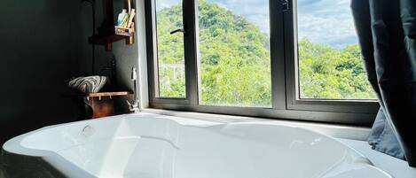 Un baño con una bañera grande, una ventana que da a vistas de follaje y un techo de madera.