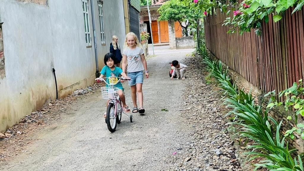A narrow alleyway with a dirt path, a child on a tricycle, and a woman walking.