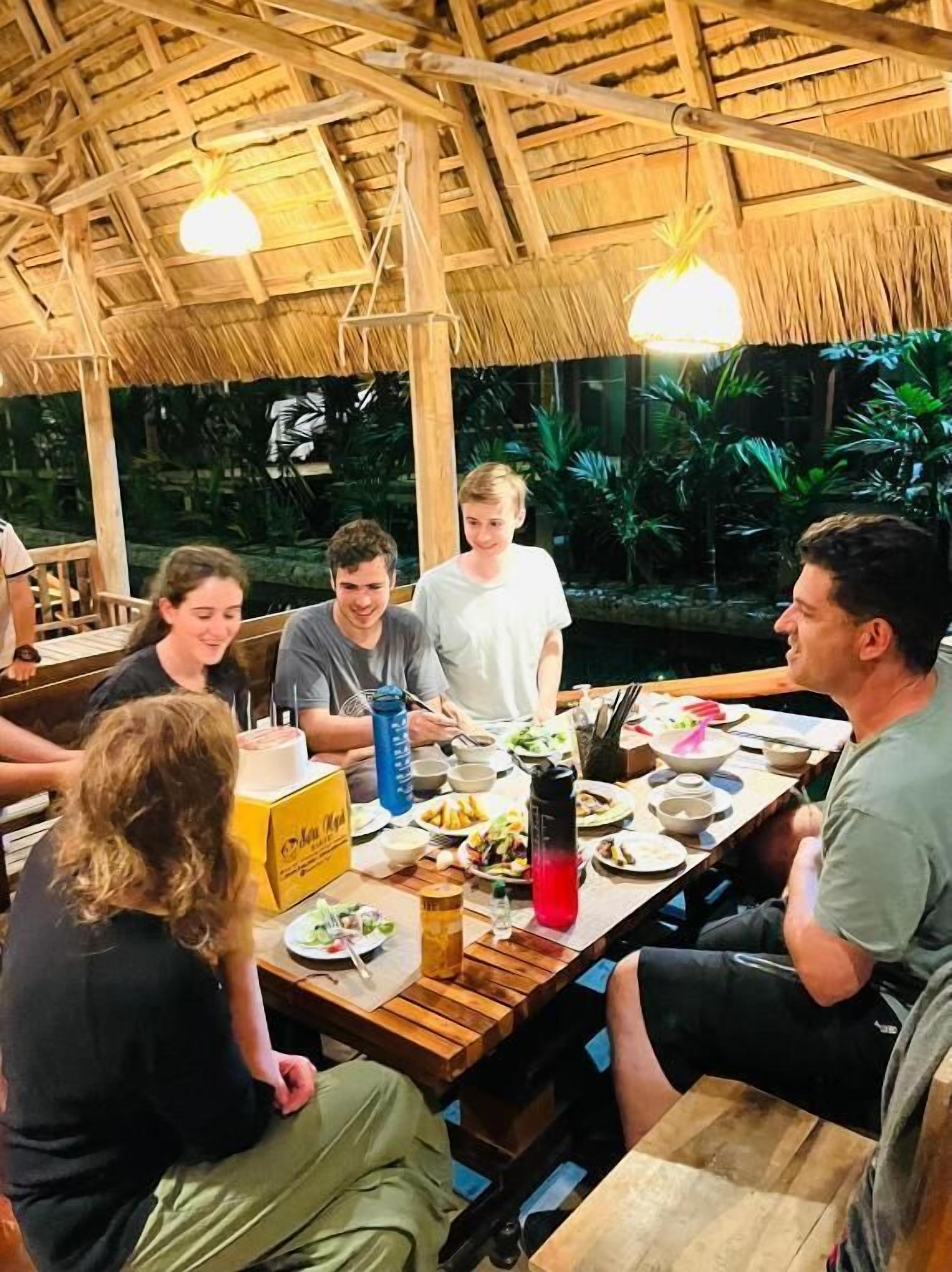 A group of people dining at a wooden table with a thatched roof, surrounded by lush greenery.