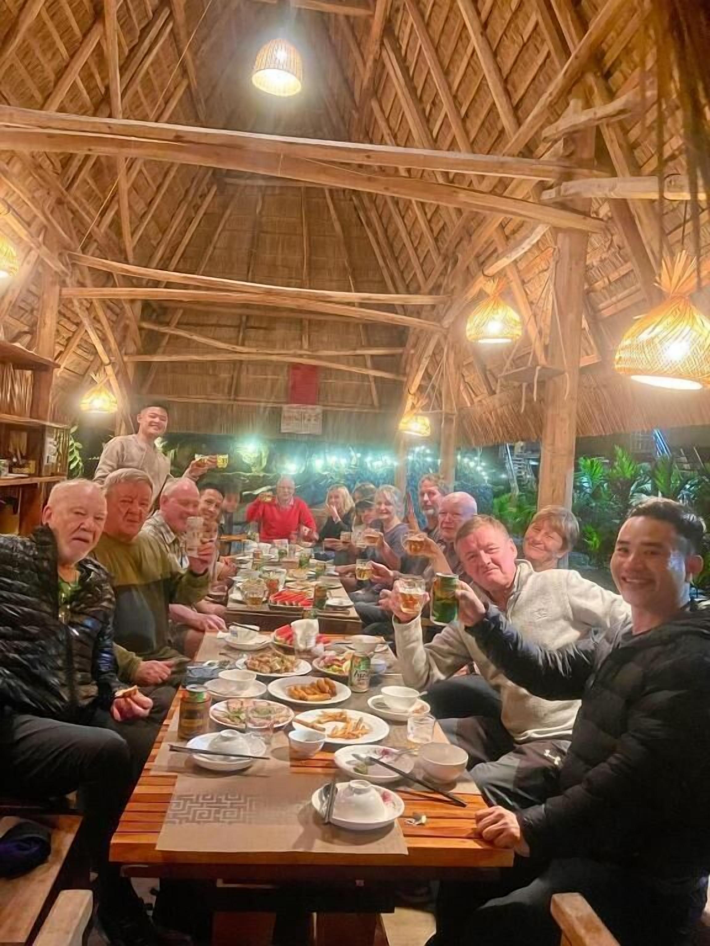 A group of people seated around a wooden table in a rustic setting with a thatched roof and hanging light fixtures.