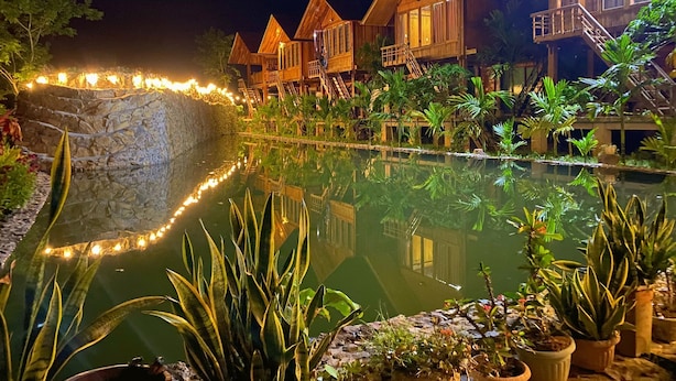 Une vue nocturne d’un complexe hôtelier avec des bâtiments en bois, un bassin décoratif et une végétation tropicale luxuriante.