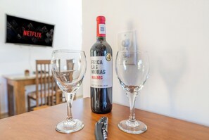 En flaske Malbec vin, to tomme vinglas, en korkskruer, et træbord, et fjernsyn med Netflix logoet, og en stol i et rum.