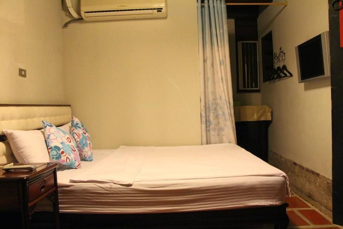Une chambre à coucher comprenant un lit, des oreillers, une table de chevet, un climatiseur, un rideau et un porte-manteau fixé au mur.